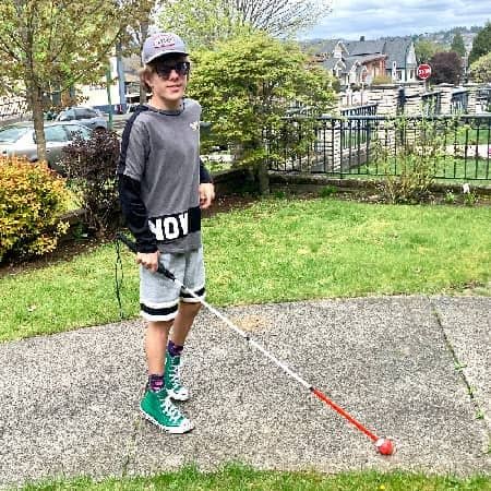 Scott in shorts, walking along on the sidewalk with his cane.